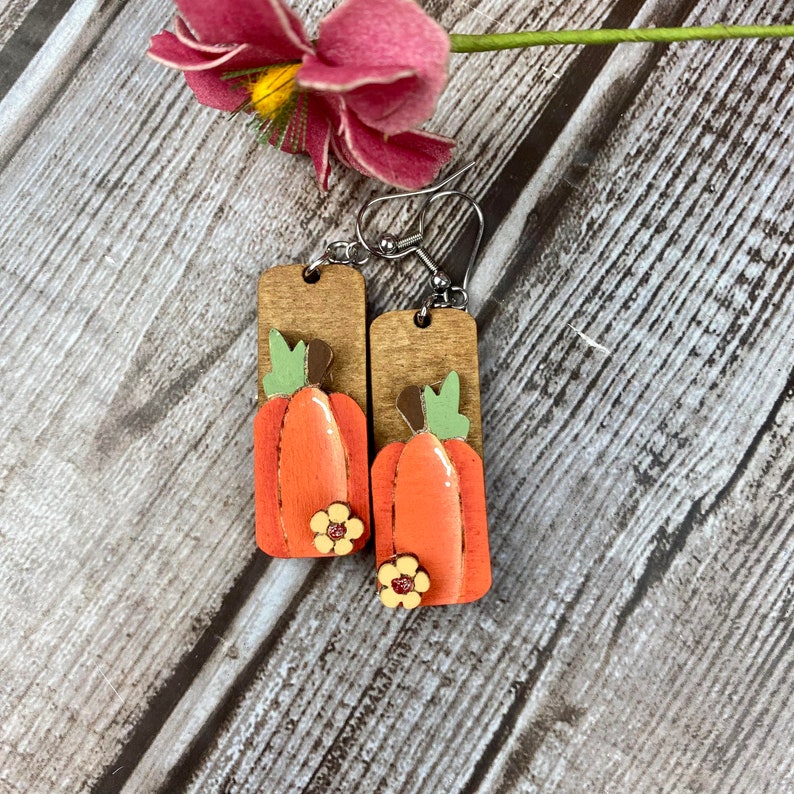Puede incluir: Un par de pendientes de madera con un dise&ntilde;o de calabaza. Las calabazas est&aacute;n pintadas de naranja con tallos verdes y detalles marrones. Cada calabaza tiene una peque&ntilde;a flor amarilla en la parte inferior.