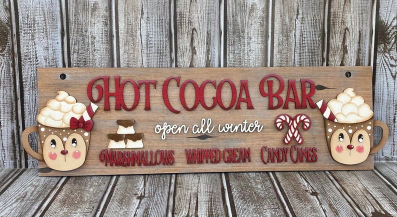 K&ouml;nnte beinhalten: Holzschilder mit dem Text "Hot Cocoa Bar" in roten Buchstaben. Das Schild sagt auch "ge&ouml;ffnet den ganzen Winter" und listet die Toppings auf: "Marshmallows", "Schlagsahne" und "Zuckerstangen". Das Schild ist mit zwei Tassen hei&szlig;er Schokolade mit Schlagsahne und einem Rentiergesicht verziert.