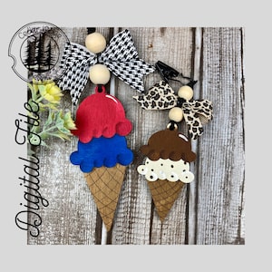 Può includere: Due decorazioni in legno a forma di cono gelato con un cono gelato rosso, bianco e blu e un cono gelato marrone, bianco e beige. I coni sono attaccati a un cordoncino nero con perline di legno e fiocchi.