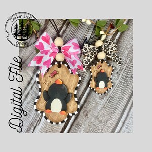 Op de afbeelding: Twee houten pinguïn-versieringen met zwart-witte strepen, een groter dan de andere. De grotere versiering heeft een roze-wit hartlint en een vlinderdetail. De kleinere versiering heeft een luipaardprintlint. Beide versieringen hebben een houten kraal aan de bovenkant.