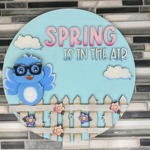 May include: A blue bird with glasses and a white belly sits on a white picket fence with pink, purple, and yellow flowers. The background is a light blue circle with white clouds and the text "SPRING IS IN THE AIR" in pink and white letters.