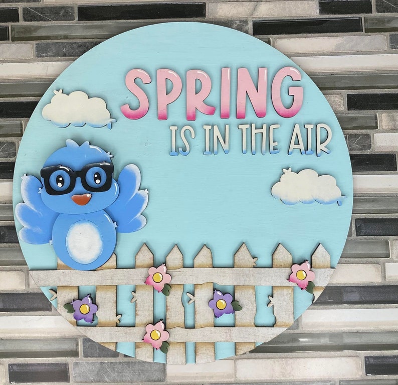 May include: A blue bird with glasses and a white belly sits on a white picket fence with pink, purple, and yellow flowers. The background is a light blue circle with white clouds and the text "SPRING IS IN THE AIR" in pink and white letters.