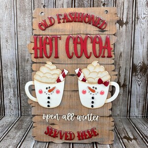 May include: A wooden sign with the text "Old Fashioned Hot Cocoa" in red letters. Two white mugs with snowman faces and red bows are on either side of the text. The sign also says "open all winter" and "served here" in red letters.