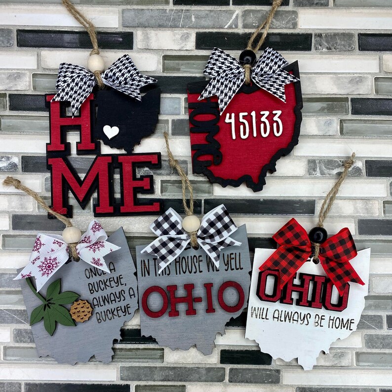 May include: Five wooden ornaments shaped like the state of Ohio. Each ornament has a different design and text. The first ornament says "Home" in red and black letters with a white heart. The second ornament is red with black trim and says "45133". The third ornament is grey with black trim and says "Once a Buckeye, Always a Buckeye". The fourth ornament is grey with black trim and says "In this house we yell Ohio". The fifth ornament is white with red trim and says "Ohio Will Always Be Home".