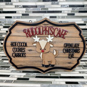 K&ouml;nnte beinhalten: Ein Schild aus Holz mit schwarzem Rand und einer Illustration von Rudolph, dem Rentier mit der roten Nase, in Rot und Wei&szlig;. Das Schild tr&auml;gt die Aufschrift "Rudolph's Cafe" und "Ge&ouml;ffnet bis sp&auml;t in die Nacht an Heiligabend". Das Schild listet auch "Hei&szlig;e Schokolade, Kekse, Karotten" auf.