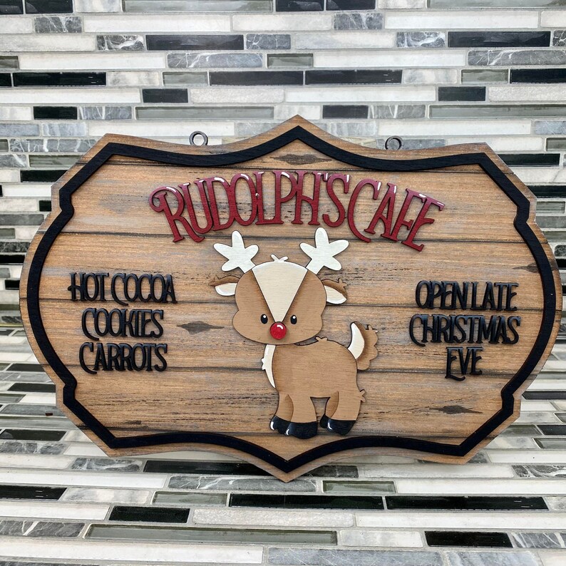 K&ouml;nnte beinhalten: Ein Schild aus Holz mit schwarzem Rand und einer Illustration von Rudolph, dem Rentier mit der roten Nase, in Rot und Wei&szlig;. Das Schild tr&auml;gt die Aufschrift "Rudolph's Cafe" und "Ge&ouml;ffnet bis sp&auml;t in die Nacht an Heiligabend". Das Schild listet auch "Hei&szlig;e Schokolade, Kekse, Karotten" auf.