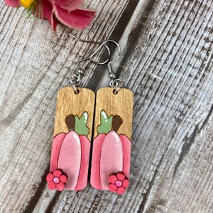Puede incluir: Un par de pendientes de madera con un dise&ntilde;o de calabaza rosa y un acento de flor rosa. Los pendientes est&aacute;n pintados con pintura marr&oacute;n, verde y rosa.