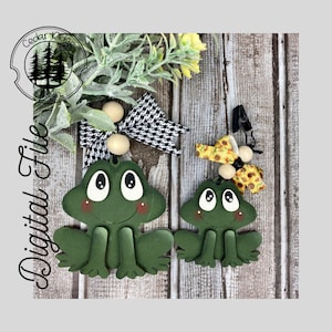 May include: Two green frog ornaments with painted eyes and smiles. The larger frog has a black and white checked bow and the smaller frog has a yellow sunflower bow.