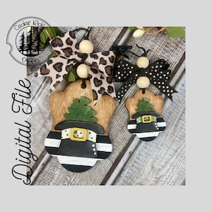 May include: Two wooden ornaments shaped like a leprechaun's hat with a green shamrock on top. The larger ornament has a black and white striped hat with a gold buckle. The smaller ornament has a black and white striped hat with a gold buckle. Both ornaments have a gold buckle and a green shamrock on top. The ornaments are hanging from a string with a wooden bead.