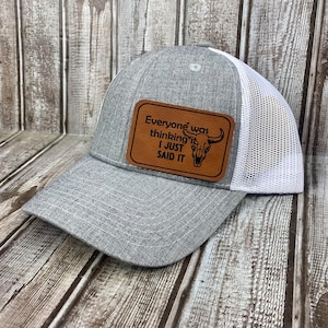 May include: Gray and white trucker hat with a brown leather patch that says "Everyone was thinking it I just said it" with a skull and horns graphic.