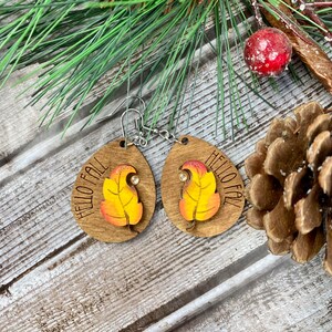May include: A pair of wooden earrings with a fall leaf design and the text "Hello Fall". The earrings are brown with a yellow and orange leaf and a small rhinestone.