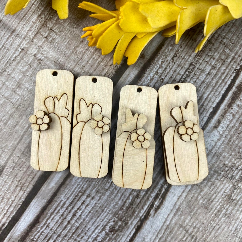Puede incluir: Cuatro recortes de madera sin terminar de calabazas con una flor en la parte superior. Los recortes son rectangulares y tienen un agujero en la parte superior para colgar.