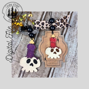 May include: Two wooden Halloween decorations featuring a skull design. One ornament has a purple candle on top and the other has a red candle. The ornament with the red candle has the text "Spooky Season" on it.