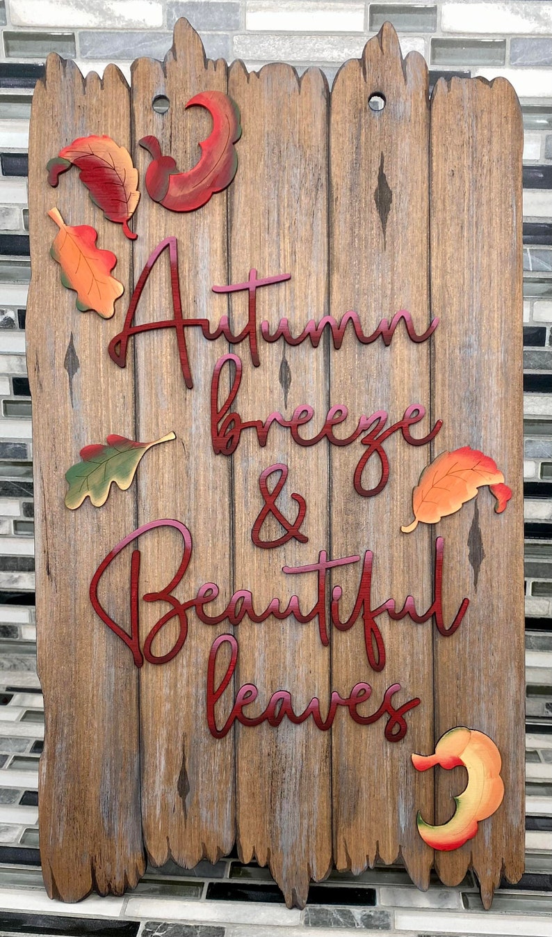 May include: Wooden sign with a rustic brown finish and red lettering that reads "Autumn breeze & Beautiful leaves". The sign is decorated with several colorful fall leaves.