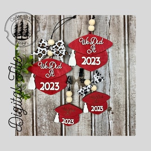 Può includere: Tre decorazioni a forma di cappello di laurea rosso con nappe bianche e il testo "We Did It 2023". Ogni decorazione è decorata con un fiocco leopardato bianco e nero e perline di legno.
