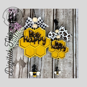 Può includere: Due ornamenti a forma di favo di miele gialli con le parole "Bee Happy" in nero. Ogni ornamento ha un cordino nero, un fiocco decorativo e un ciondolo a forma di ape. Un fiocco è a scacchi bianchi e neri, l'altro è leopardato.