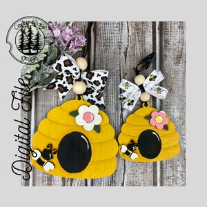 May include: Two wooden beehive ornaments with a black bee and a pink flower on each. The larger beehive has a leopard print bow and wooden beads. The smaller beehive has a white bow with bees printed on it and wooden beads.