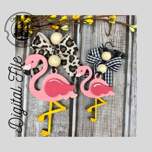 May include: Two pink wooden flamingo ornaments with yellow legs and black eyes. The larger flamingo has a leopard print bow and the smaller flamingo has a black and white checked bow. Both ornaments have wooden beads and a clip for hanging.