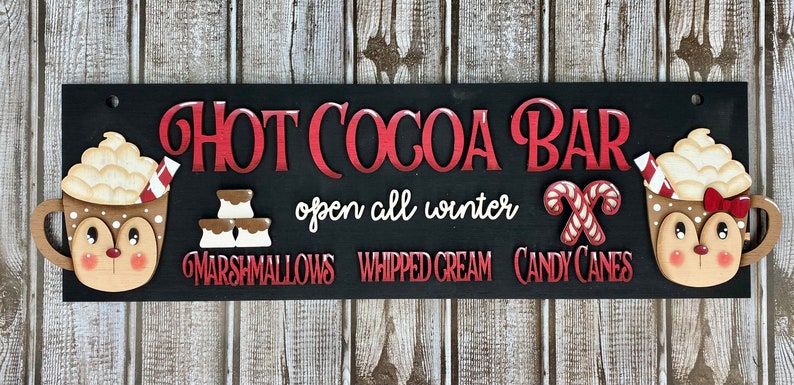 K&ouml;nnte beinhalten: Ein schwarzes Holzschild mit dem Text "HOT COCOA BAR" in roten Buchstaben. Das Schild enth&auml;lt auch den Text "open all winter" und Bilder einer Tasse hei&szlig;er Schokolade mit Schlagsahne und Zuckerstangen. Der Text "Marshmallows", "Schlagsahne" und "Zuckerstangen" ist ebenfalls auf dem Schild enthalten.