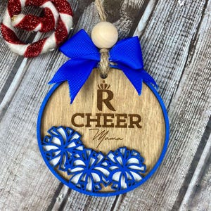Op de afbeelding: Een houten kerstversiering met een blauw lint en pompoenen. De versiering is versierd met een cheerleading pompon-ontwerp en de tekst "R Cheer Mama".