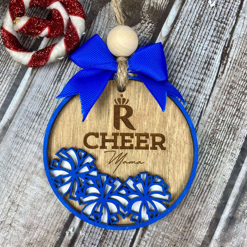 Op de afbeelding: Een houten kerstversiering met een blauw lint en pompoenen. De versiering is versierd met een cheerleading pompon-ontwerp en de tekst "R Cheer Mama".