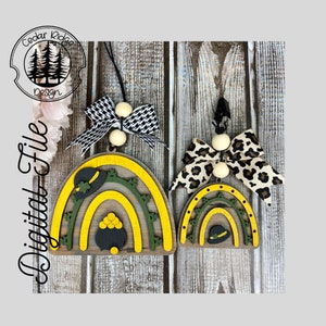 Puede incluir: Dos adornos de arcoíris de madera con un pote de oro y un sombrero de duende. El arcoíris más grande es amarillo y verde con un pote de oro negro. El arcoíris más pequeño es amarillo y verde con un sombrero de duende negro. Ambos arcoíris tienen un lazo blanco con cuentas.