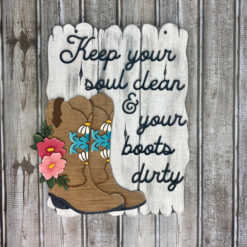 May include: Wooden sign with a whitewashed background and black lettering that reads "Keep your soul clean & your boots dirty". Two brown cowboy boots with blue and white floral designs are attached to the sign.