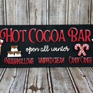 K&ouml;nnte beinhalten: Ein schwarzes Holzschild mit dem Text "Hot Cocoa Bar" in roten Buchstaben. Das Schild enth&auml;lt auch den Text "open all winter" und listet die Zutaten auf: "Marshmallows", "Schlagsahne" und "Zuckerstangen". Das Schild ist mit zwei Rentierbechern mit Schlagsahne und Zuckerstangen verziert.