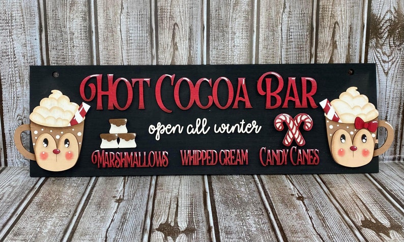 K&ouml;nnte beinhalten: Ein schwarzes Holzschild mit dem Text "Hot Cocoa Bar" in roten Buchstaben. Das Schild enth&auml;lt auch den Text "open all winter" und listet die Zutaten auf: "Marshmallows", "Schlagsahne" und "Zuckerstangen". Das Schild ist mit zwei Rentierbechern mit Schlagsahne und Zuckerstangen verziert.