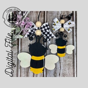 May include: Two wooden bee ornaments painted black and yellow, one larger than the other. The larger bee has white glitter wings and is decorated with a black and white checked ribbon and wooden beads. The smaller bee has white wings and is decorated with a white ribbon with a bee pattern.
