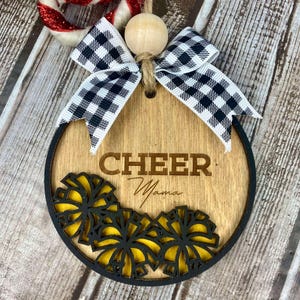 Op de afbeelding: Een ronde houten ornament met een zwart-wit geblokt lint, een houten kraal en de tekst "CHEER Mama" gegraveerd op de voorkant. De ornament is versierd met gele en zwarte cheerleading pompoenen.