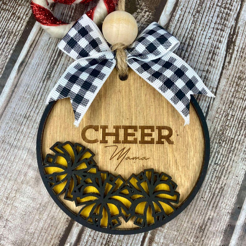 Op de afbeelding: Een ronde houten ornament met een zwart-wit geblokt lint, een houten kraal en de tekst "CHEER Mama" gegraveerd op de voorkant. De ornament is versierd met gele en zwarte cheerleading pompoenen.