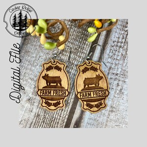 May include: A pair of wooden earrings with a pig silhouette and the text "Farm Fresh". The earrings are brown and have a rustic look.