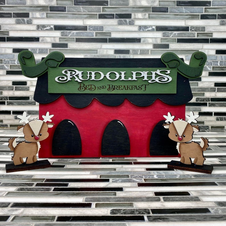 Op de afbeelding: Een rood houten bord met een zwart dak en groene versieringen. Het bord heeft de tekst "Rudolph's Bed and Breakfast" in wit cursief schrift. Twee houten rendierfiguren staan aan weerszijden van het bord.