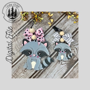 Può includere: Due ornamenti di procione in legno con fiocchi rosa e grigi. Il procione più grande ha un fiocco rosa con macchie nere e il procione più piccolo ha un fiocco grigio con strisce bianche. Entrambi gli ornamenti hanno occhi neri e un naso rosa.