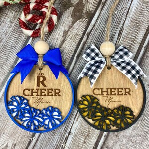 Op de afbeelding: Twee houten kerstversieringen met een cheer-thema. Een versiering is blauw met een blauw-witte pompon en de tekst "R Cheer Mama". De andere versiering is zwart-wit met een zwart-gele pompon en de tekst "Cheer Mama".