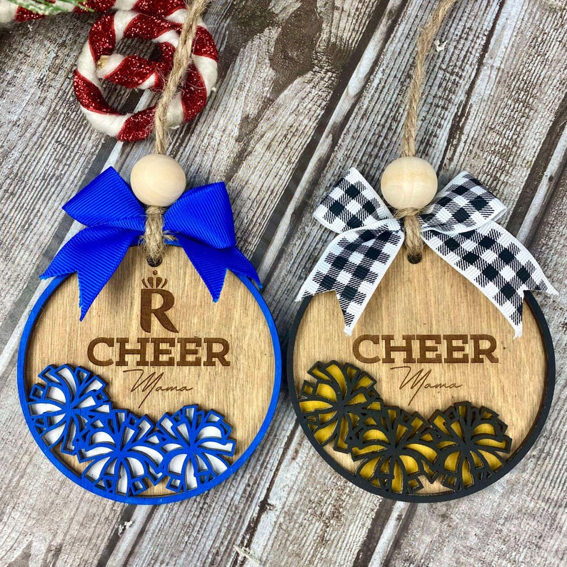 Op de afbeelding: Twee houten kerstversieringen met een cheer-thema. Een versiering is blauw met een blauw-witte pompon en de tekst "R Cheer Mama". De andere versiering is zwart-wit met een zwart-gele pompon en de tekst "Cheer Mama".