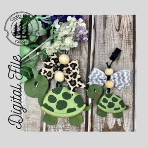 Könnte beinhalten: Zwei hölzerne Schildkröten-Anhänger, die in Grün und Braun bemalt sind. Die größere Schildkröte hat eine Schleife mit Leopardenmuster und die kleinere Schildkröte hat eine Schleife mit grau-weißem Chevron-Muster. Beide Schildkröten haben Holzperlen und einen schwarzen Clip.