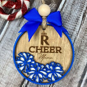 Op de afbeelding: Een houten kerstversiering met een blauw lint en een kroon met de letter R. De versiering is versierd met witte en blauwe pompoenen en de woorden "Cheer Mama" zijn in het hout gegraveerd.