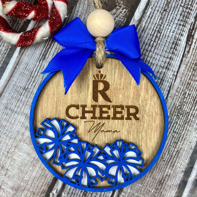 Op de afbeelding: Een houten kerstversiering met een blauw lint en een kroon met de letter R. De versiering is versierd met witte en blauwe pompoenen en de woorden "Cheer Mama" zijn in het hout gegraveerd.