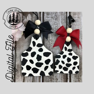 Pode incluir: Duas etiquetas de madeira com estampa de vaca com laços pretos e vermelhos e contas de madeira. As etiquetas estão penduradas em uma superfície de madeira.