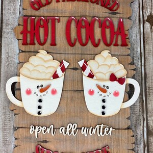 May include: Wooden sign with the text "Old Fashioned Hot Cocoa" in red letters. Two white mugs with snowman faces and red and white candy canes are on either side of the text. The sign also says "open all winter" and "SERVED HERE" in red letters.