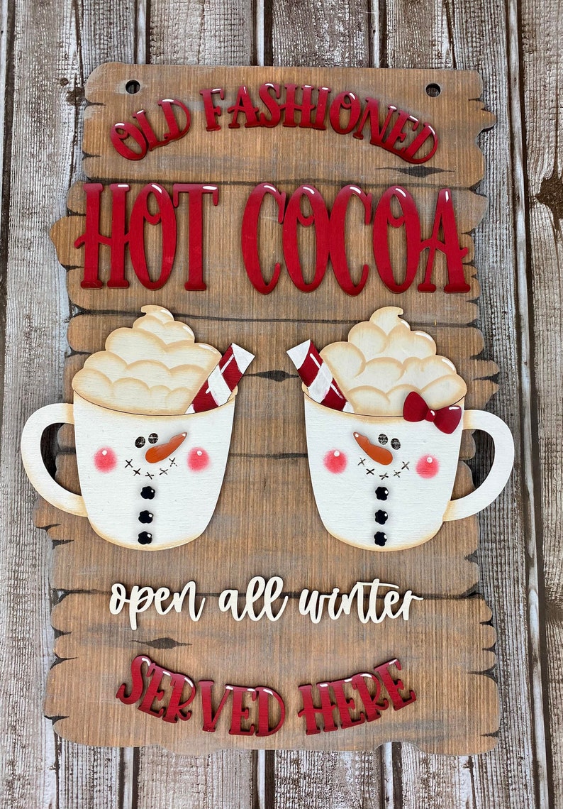May include: Wooden sign with the text "Old Fashioned Hot Cocoa" in red letters. Two white mugs with snowman faces and red and white candy canes are on either side of the text. The sign also says "open all winter" and "SERVED HERE" in red letters.