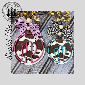 Può includere: Due ornamenti rotondi in legno con un design a stampa di mucca. Un ornamento ha un contorno di cactus rosa e l'altro ha un contorno di cactus turchese. Entrambi gli ornamenti hanno un centro in legno marrone e un anello esterno bianco. Ogni ornamento ha un fiocco a stampa leopardata con una perla di legno.