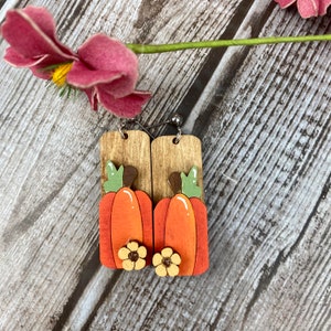 Puede incluir: Un par de pendientes de calabaza de madera con fondo de madera marr&oacute;n y tallo verde. Las calabazas est&aacute;n pintadas de naranja con una flor amarilla en la parte inferior.