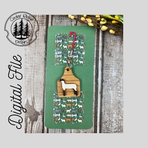 May include: A green card with a white lamb cutout on a wooden tag hanging from a red and white string. The card has a pocket with a green and white farm animal print. The text "Cedar Ridge Design" is on the top left corner of the card.