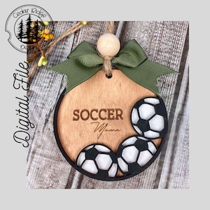 May include: A wooden ornament with a green bow and three soccer balls. The ornament is engraved with the words "Soccer Mama".