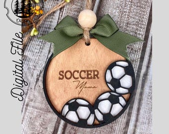 Könnte beinhalten: Ein Holz-Anhänger mit einer grünen Schleife und drei Fußbällen. Der Anhänger ist mit den Worten "Soccer Mama" graviert.