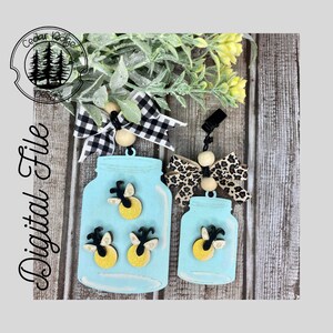 May include: Two wooden mason jar ornaments with fireflies inside. The larger jar has three fireflies and the smaller jar has one firefly. Both jars are painted a light blue color. The jars are hanging from black clips with a bow and wooden beads.
