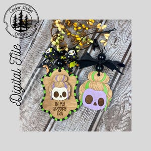 Op de afbeelding: Twee houten Halloween-ornamenten. Eén heeft een schedel met een knot en de tekst "IN MY SPOOKY ERA". De andere heeft een paarse schedel met groen haar. Beide zijn versierd met zwarte strikken en decoratieve accenten.